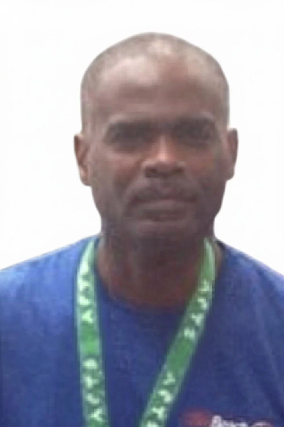 A man with a shaved head, wearing a blue shirt and a green lanyard around his neck, posing against a plain white background.