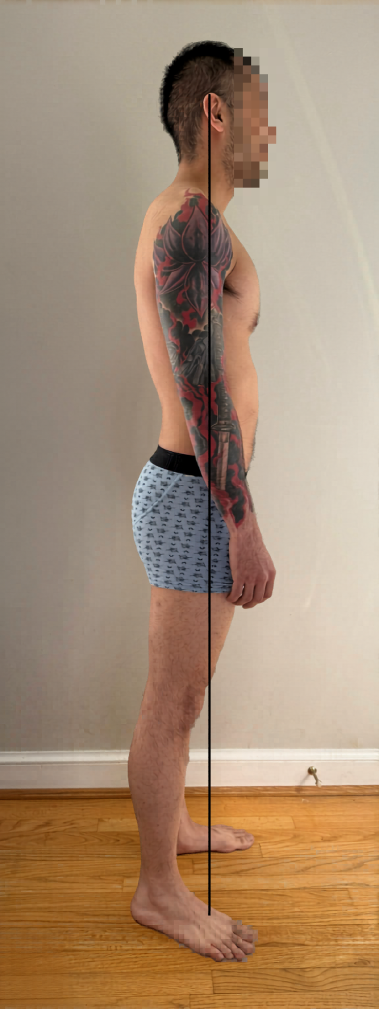 Side view of a shirtless man with tattoos, wearing boxer shorts, standing on a wooden floor against a plain wall.