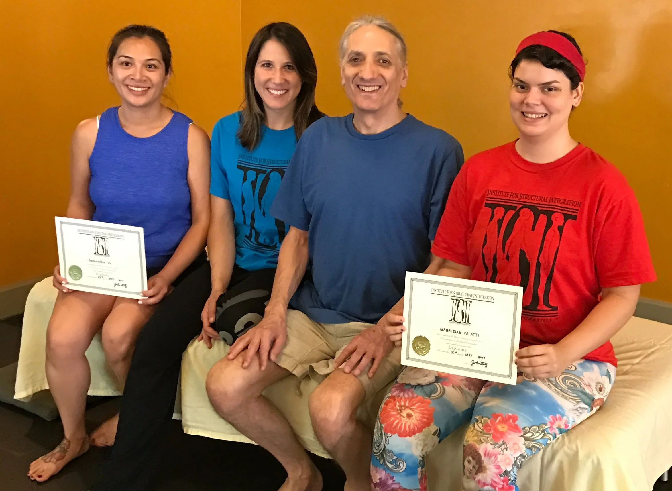 Four people sitting on a yellow sofa holding certificates, smiling at the camera. The background is a solid yellow wall.