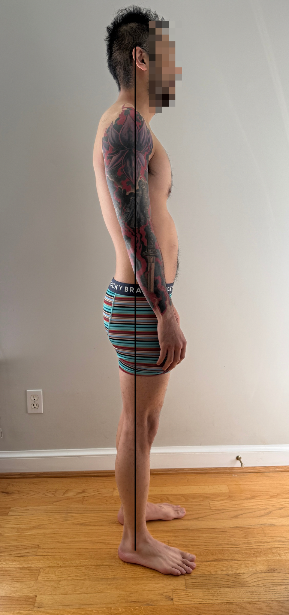 Side view of a shirtless man with a large tattoo on his left arm, standing against a plain gray wall with hardwood flooring, wearing striped boxer shorts, hands at his side, with his face pixelated.