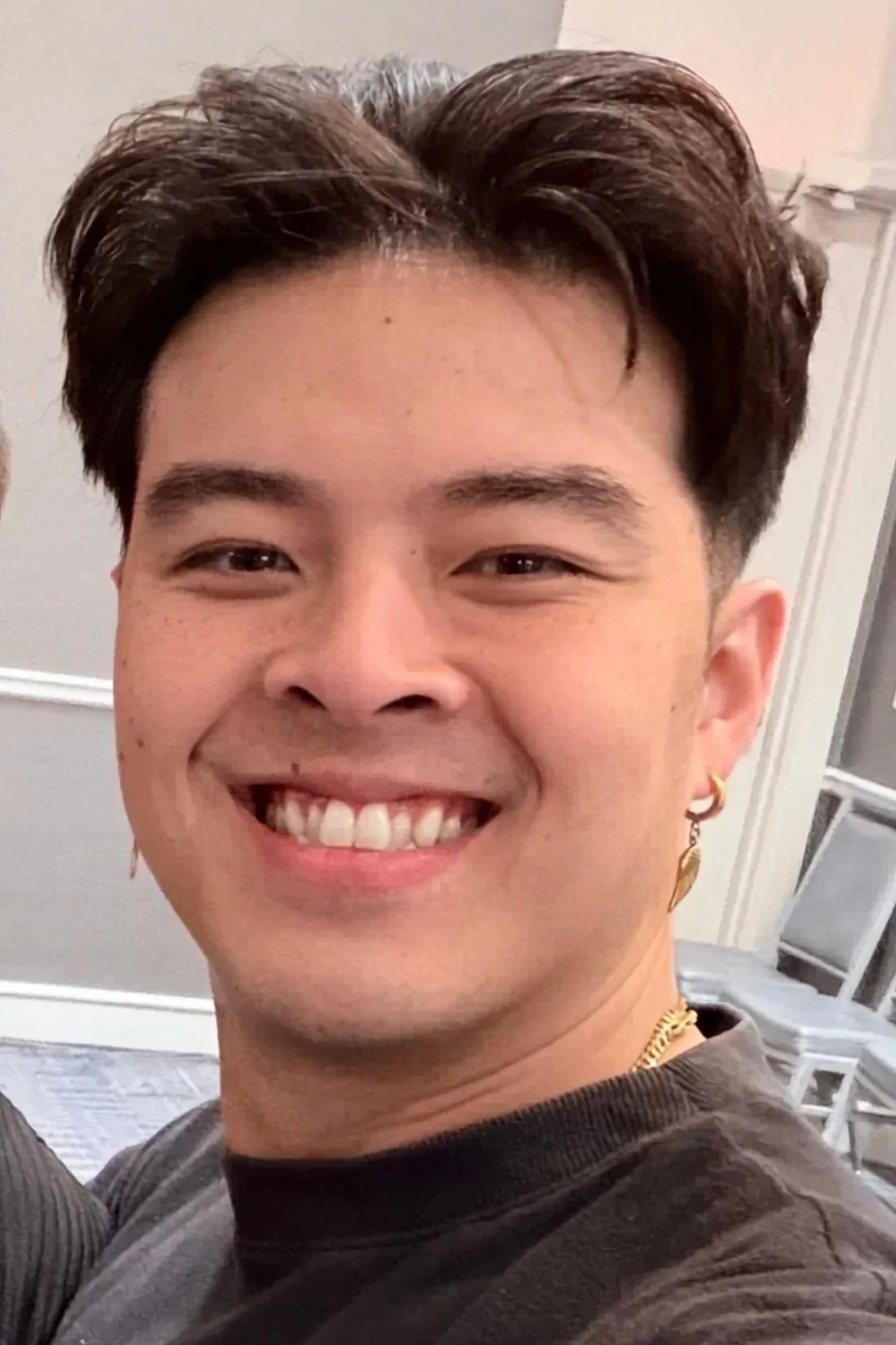 A young person with short dark hair, wearing earrings and a necklace, smiling in front of a plain wall and some furniture.