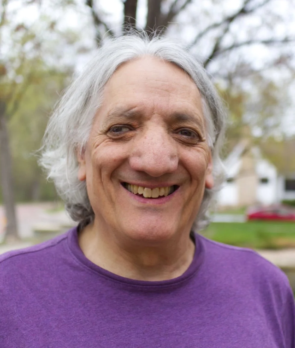 A joyful man with long gray hair wearing a purple shirt standing outdoors with trees and houses in the background.