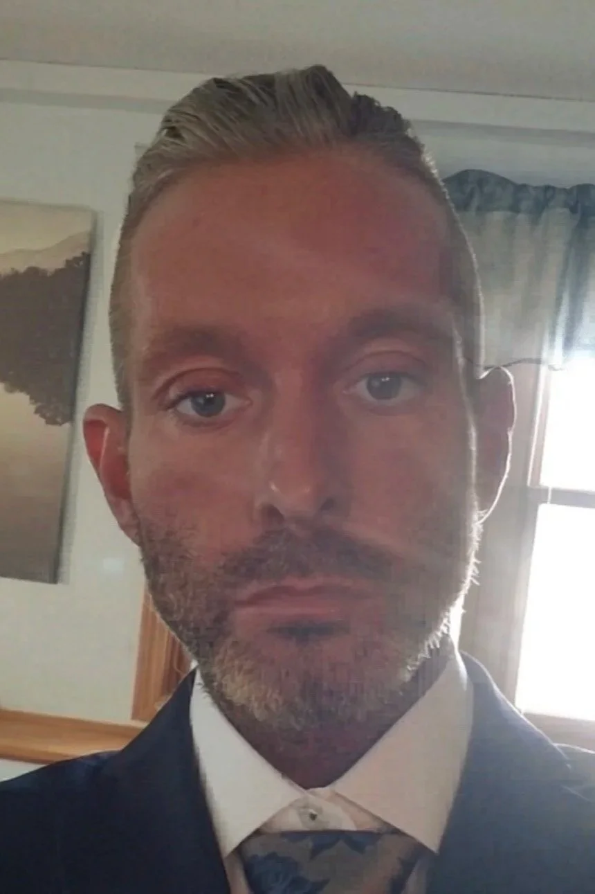 A man with short gray hair and a beard, wearing a suit and tie, taking a selfie indoors near a window with curtains.