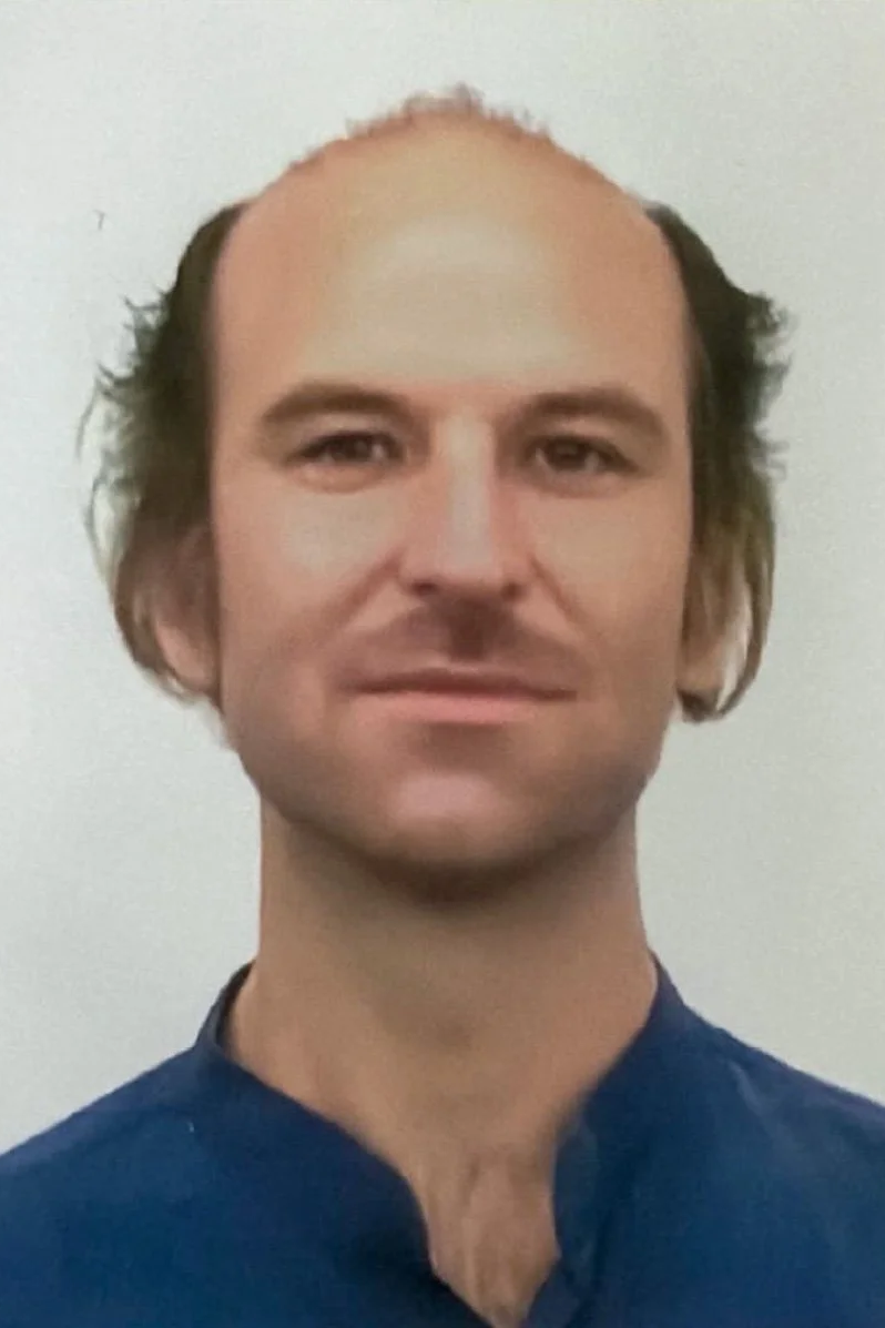 A man with receding hairline and medium-length brown hair, wearing a dark blue shirt, looking at the camera with a neutral expression, standing against a plain light-colored background.