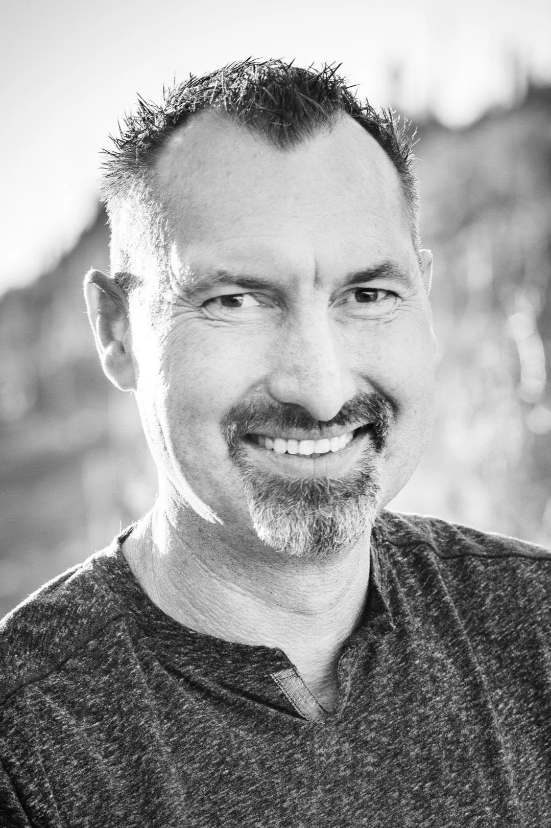 Black and white photo of a smiling man with short hair, mustache, and goatee, wearing a dark sweater, outdoors with blurred background.