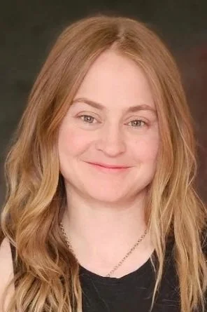 A young woman with long, wavy red hair smiling at the camera, wearing a black top and a necklace.