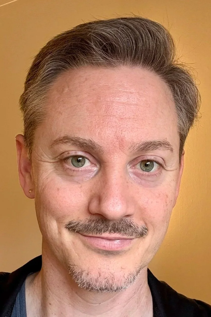 Close-up of a smiling man with short, slicked-back hair, light skin, a goatee, and light green eyes, wearing a black shirt, against a yellow background.