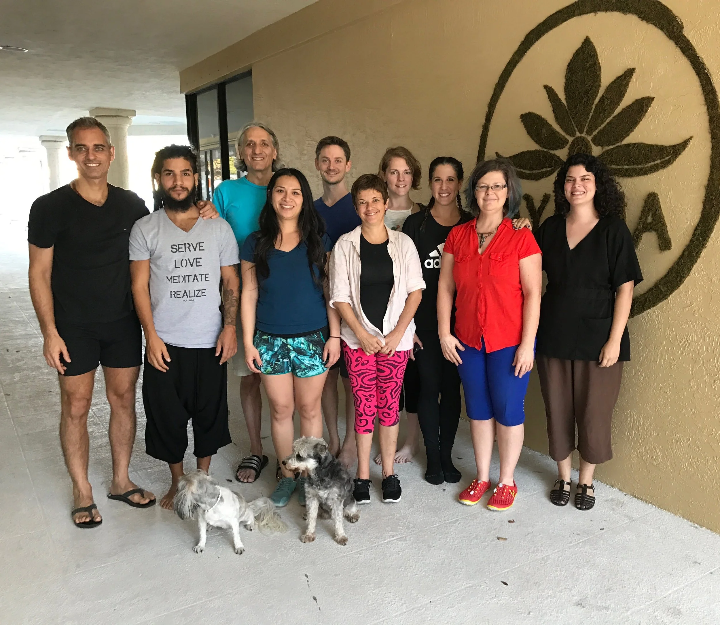 Group of ten diverse people posing indoors with two dogs in front of a wall with a large botanical design and the word 'YOGA' partially visible. The individuals are smiling and dressed casually.