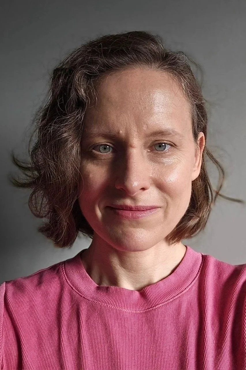 Close-up of a woman with short, wavy brown hair wearing a pink shirt, looking at the camera with a neutral expression, against a plain gray background.