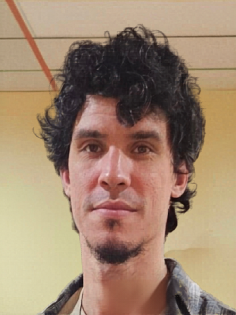 A man with curly black hair, light skin, and a small goatee, looking directly at the camera. He is inside a room with a yellow wall and some visible ceiling lines.