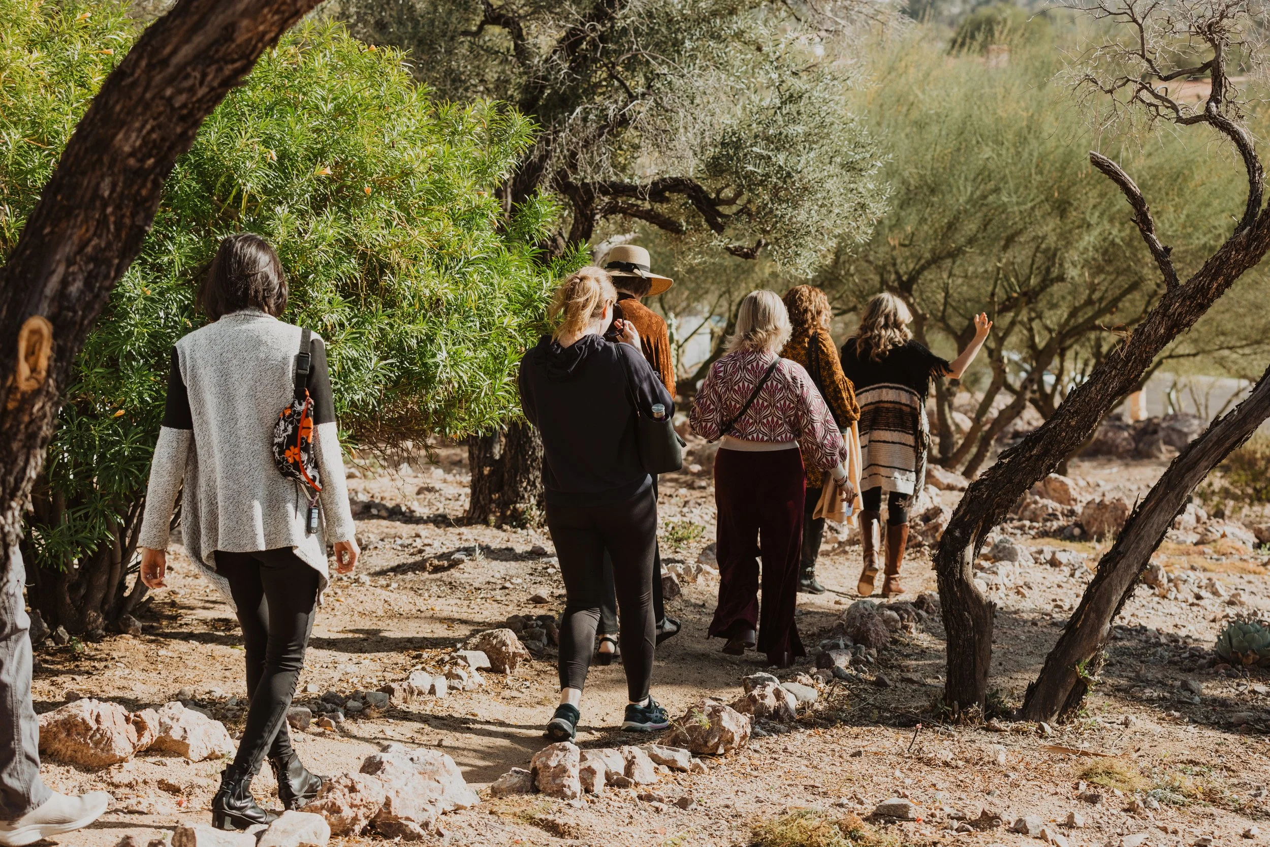 A group of people walking along a rocky dirt path surrounded by trees and green bushes in a natural outdoor setting.