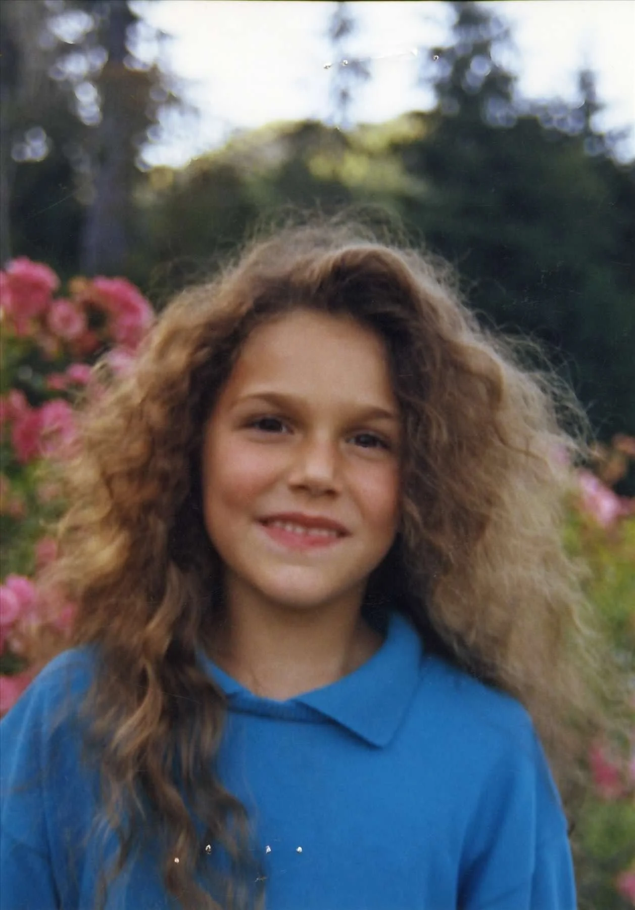 A young girl with curly hair wearing a blue hoodie outdoors, surrounded by pink flowers and greenery, smiling at the camera.