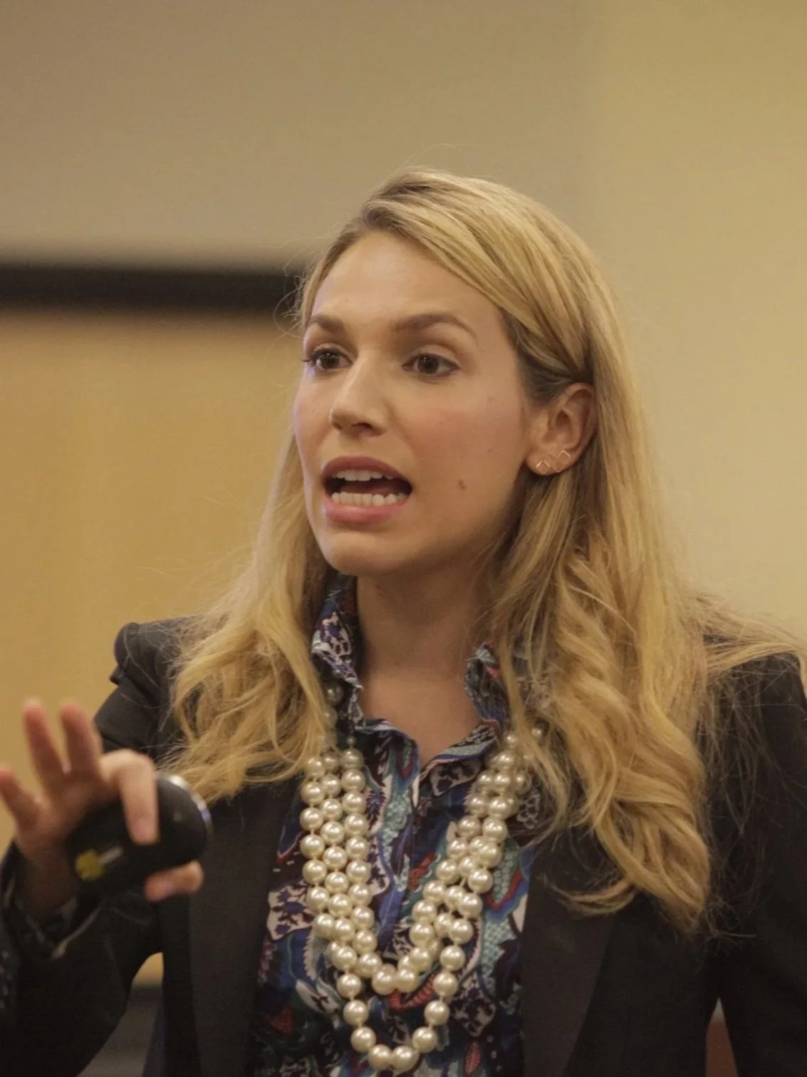 A woman with long blonde hair, wearing a blazer and pearl necklace, speaking and gesturing during a presentation or speech.