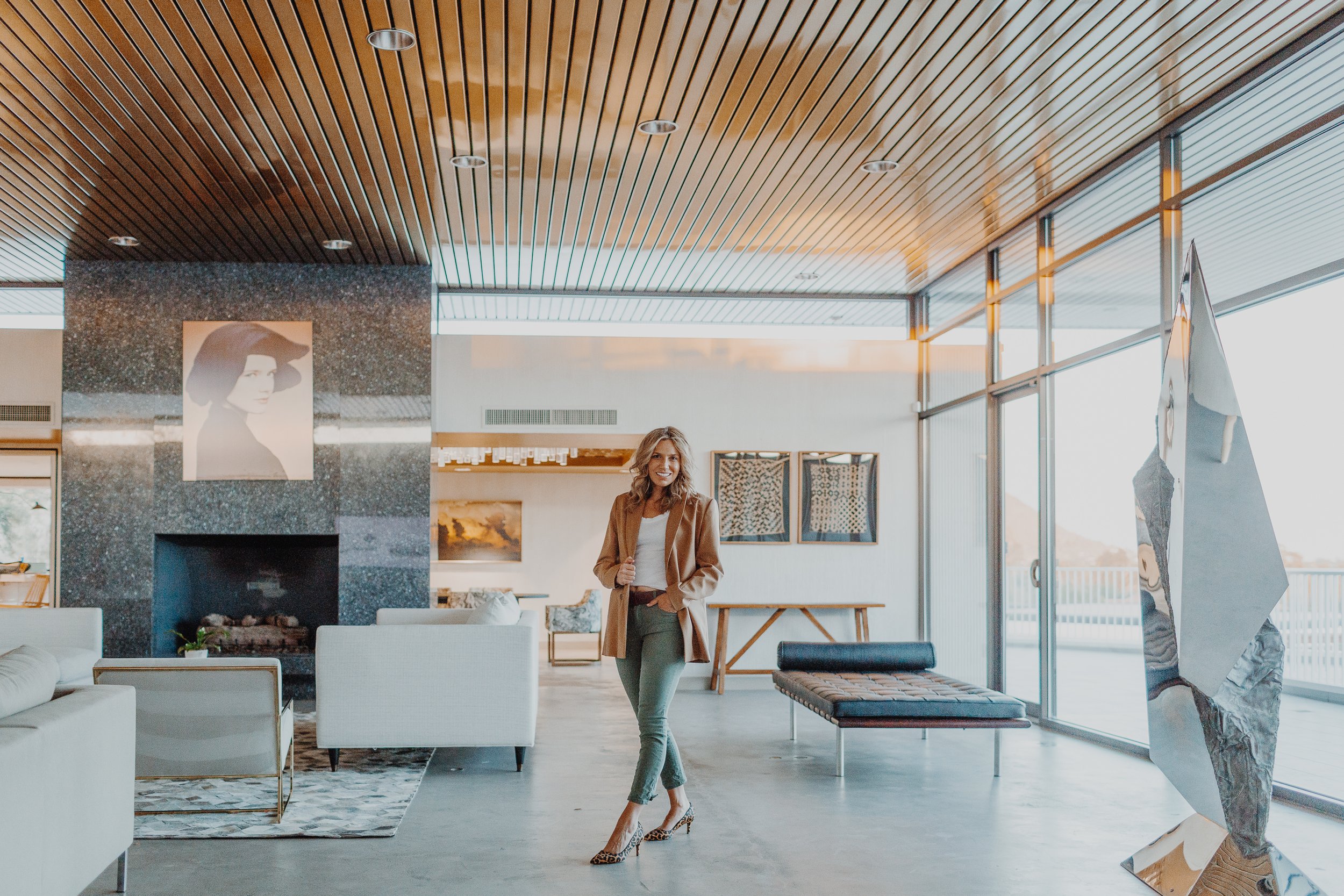Woman standing in a modern art gallery with artwork and sculptures, surrounded by large windows and a wood-paneled ceiling.