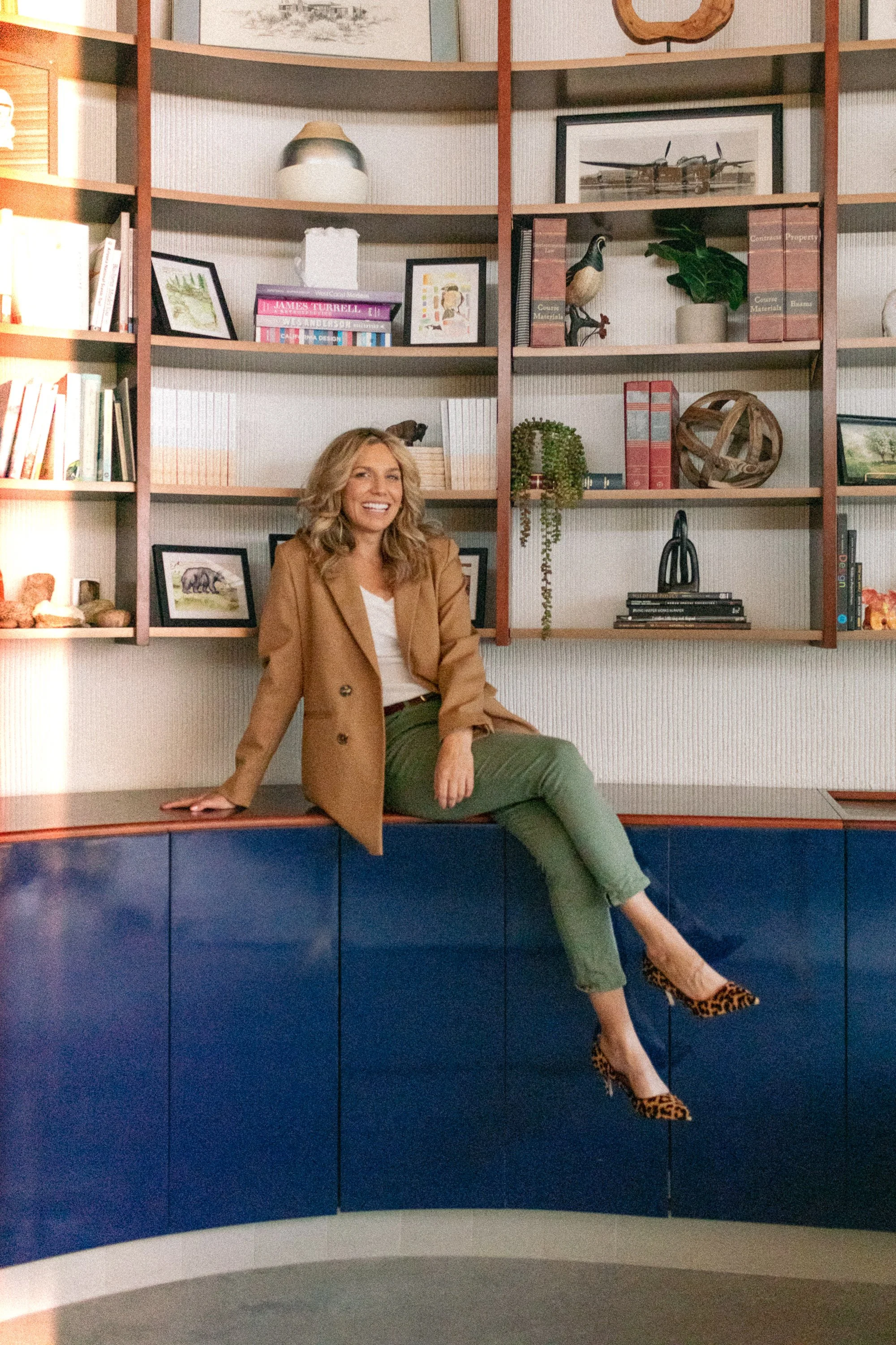 Woman sitting on a cabinet on a modern, decorated bookshelf wall in a stylish room. She is smiling, wearing a beige blazer, white top, green pants, and leopard print high heels.