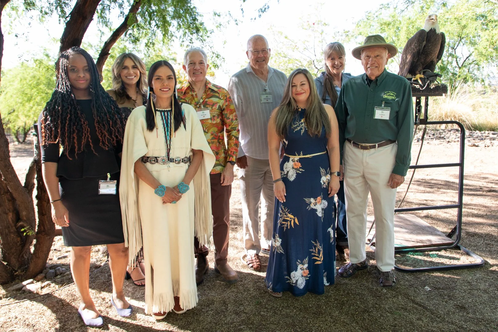 Group of nine people standing outdoors with trees and a bald eagle on a stand in the background.