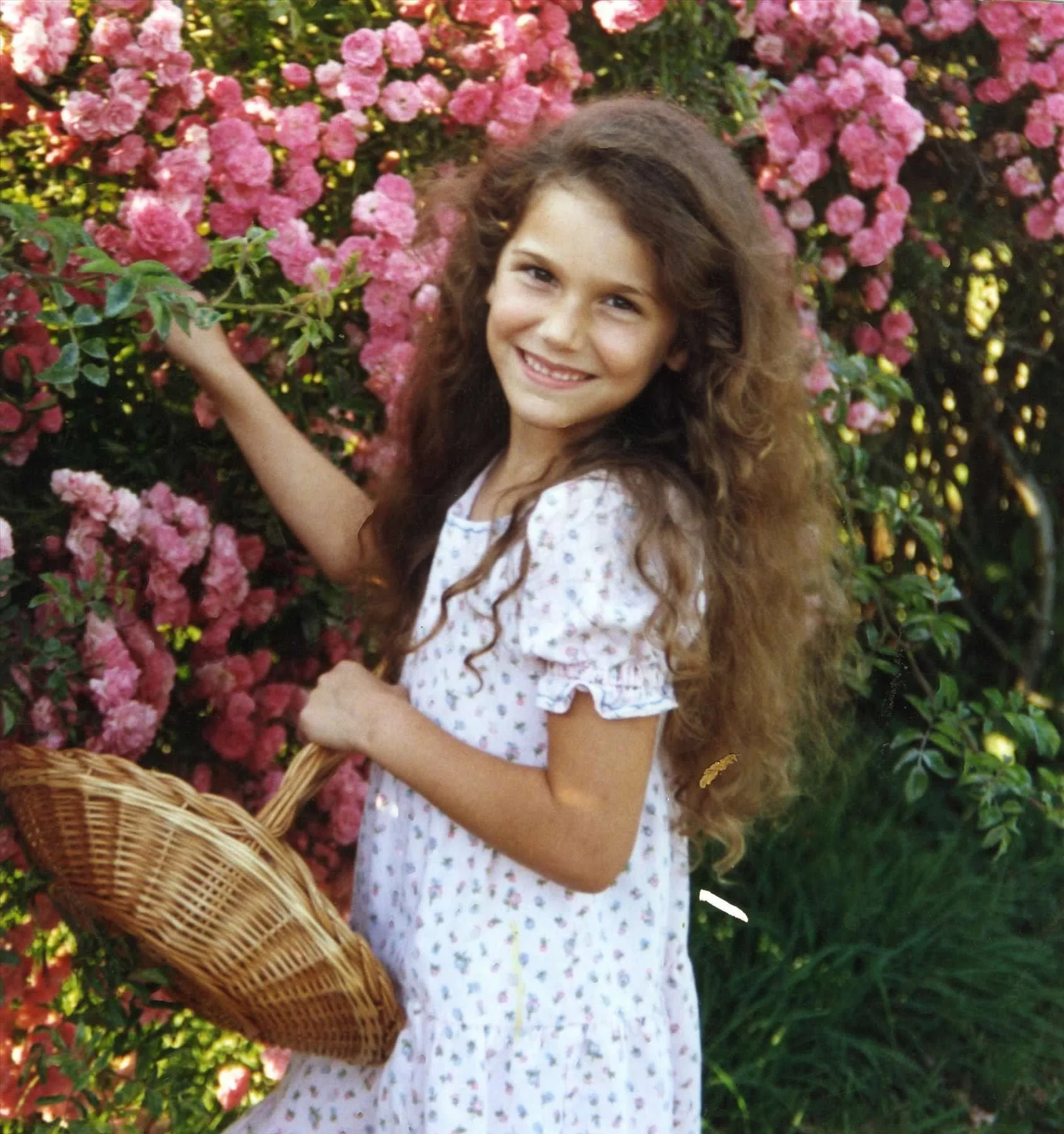 Young girl with long curly hair smiling while holding a wicker basket, standing next to a bush with pink flowers.