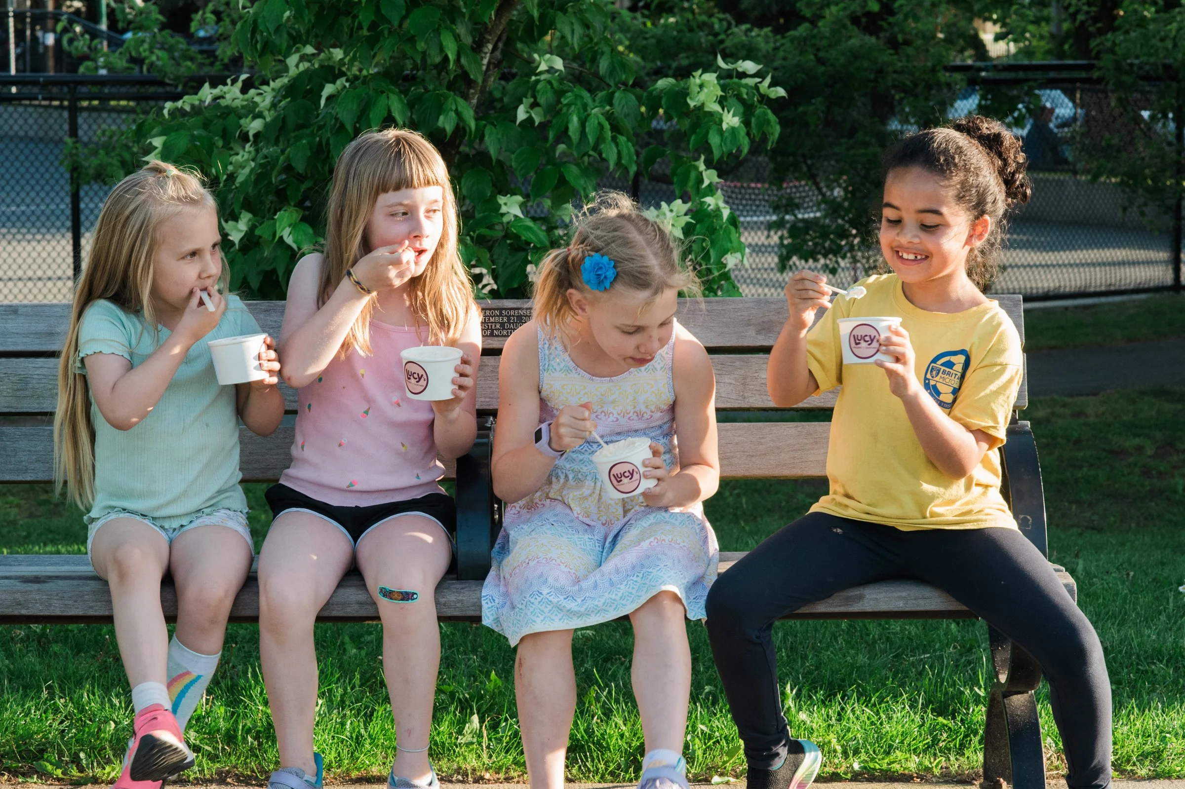 Five young girls sitting on a park bench enjoying ice cream, with green trees and a fence in the background.