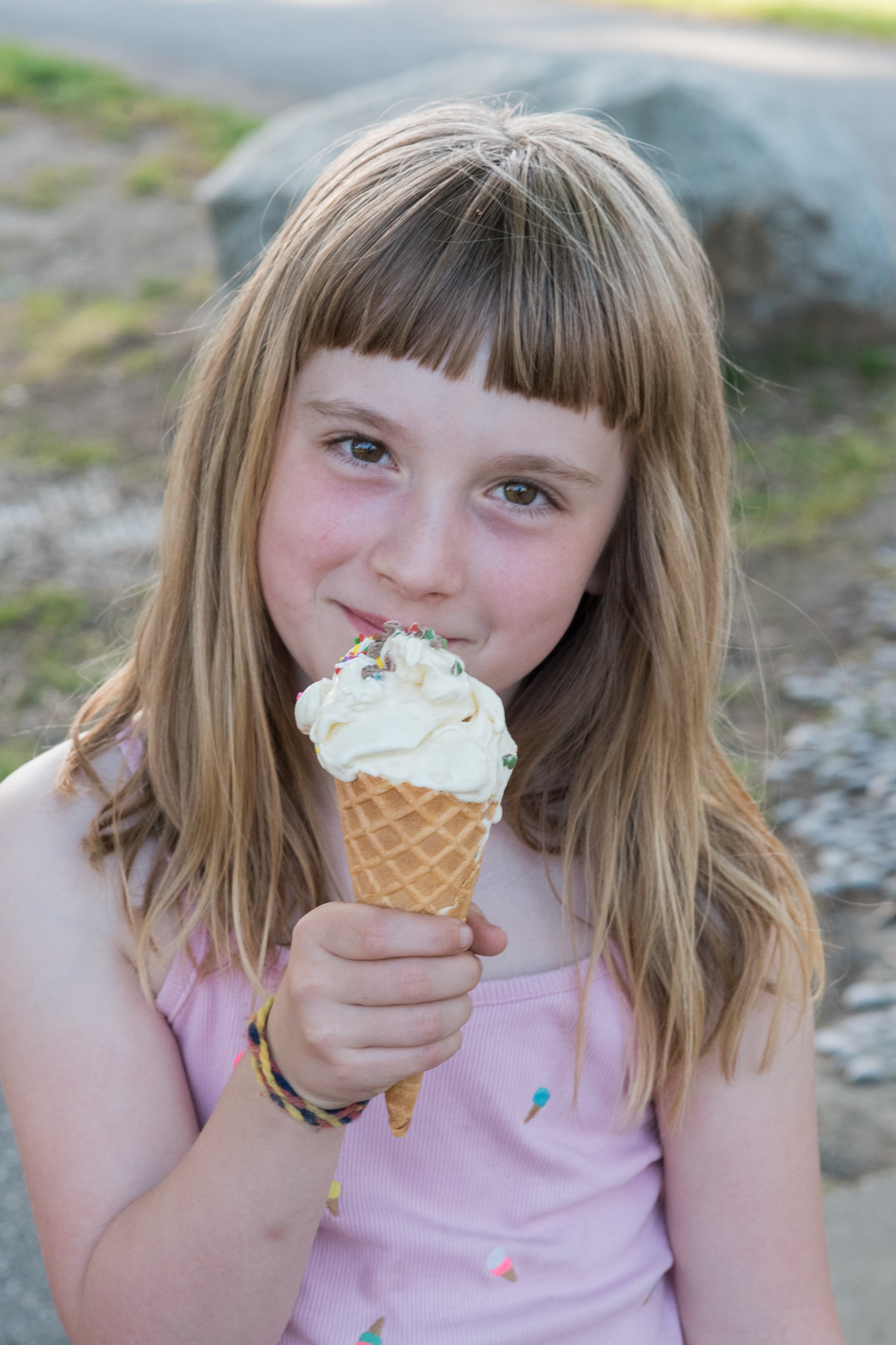 A young girl with long, light brown hair and a pink tank top, holding a vanilla ice cream cone with rainbow sprinkles, Frozen Custard East Vancouver, BC