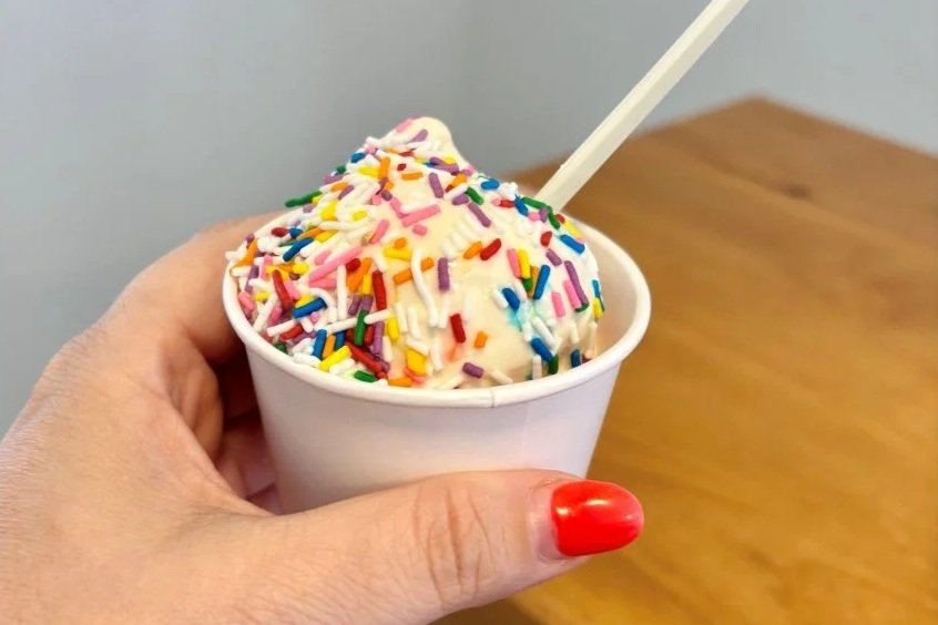 A hand holding a small white cup of vanilla ice cream topped with rainbow sprinkles, with a white spoon in the ice cream. Frozen Custard East Vancouver, BC