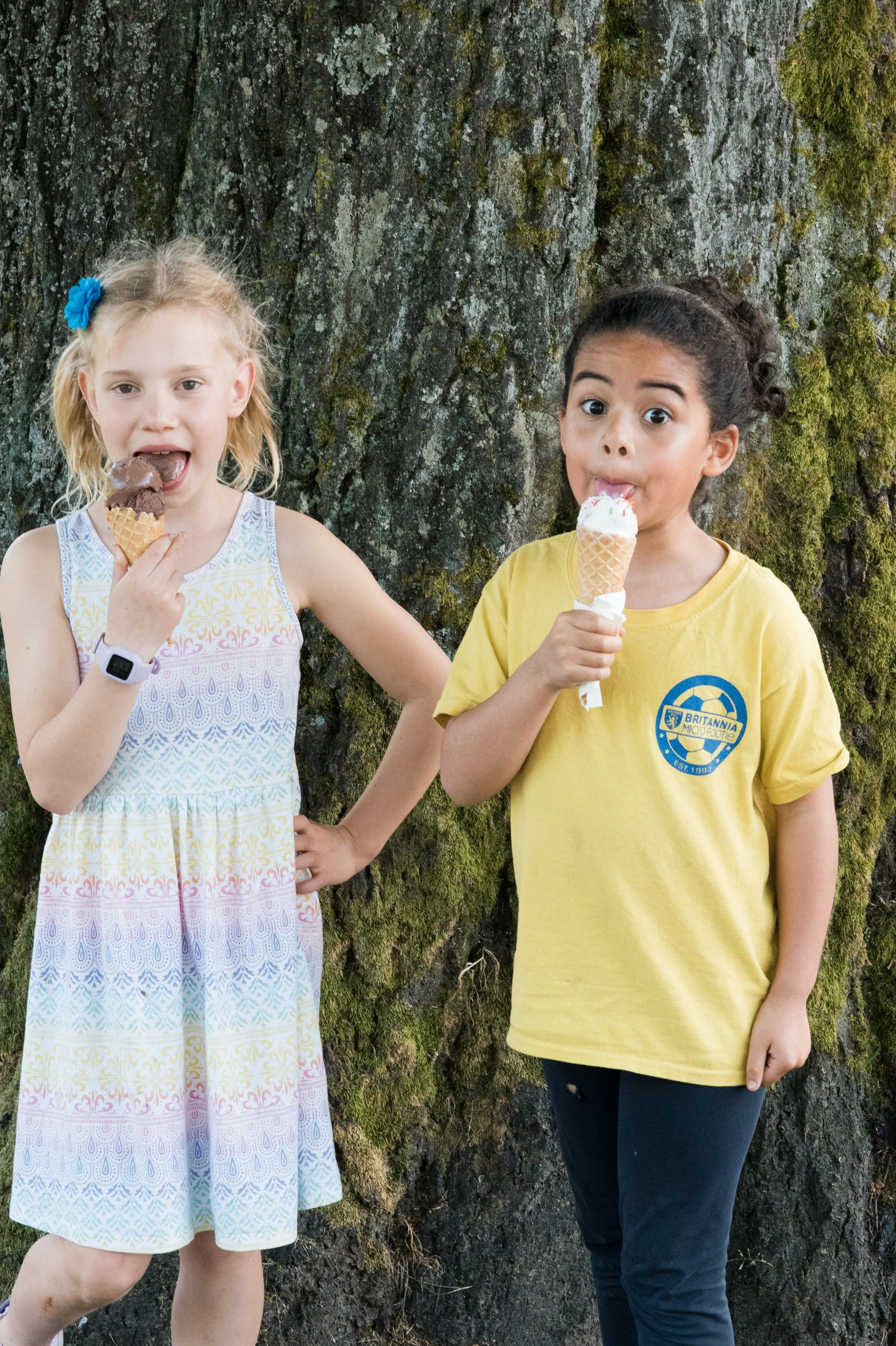 Two young girls standing in front of a moss-covered tree, each holding and eating ice cream cones, one with vanilla and sprinkles and the other with chocolate. Frozen Custard East Vancouver, BC