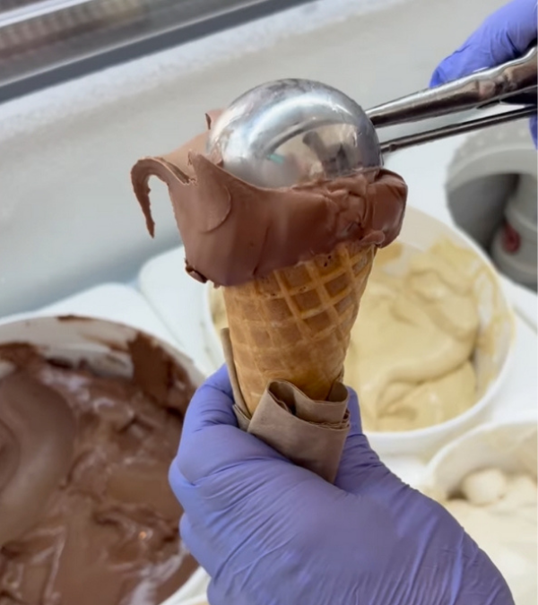 A person wearing purple gloves scoops chocolate ice cream onto a cone using a metal ice cream scoop, with vanilla ice cream in the background. Frozen Custard East Vancouver, BC