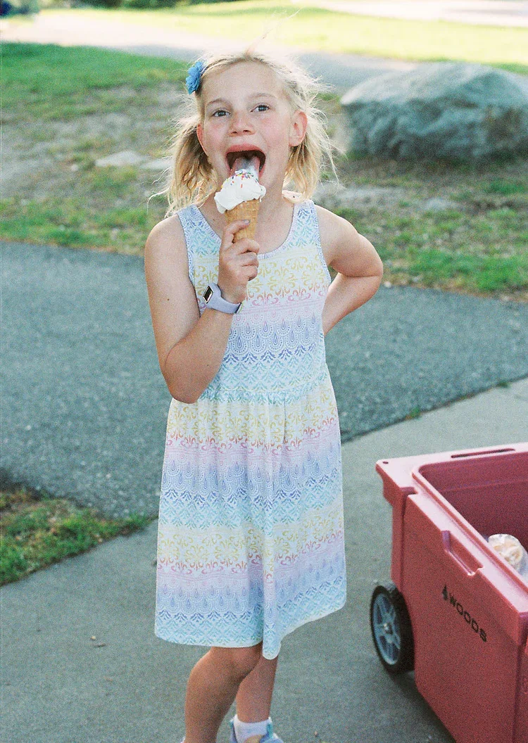 A young girl in a sleeveless pastel-colored dress with a rainbow pattern, holding an ice cream cone, smiling with her mouth open, outside on a sidewalk with a red wagon nearby. Frozen Custard East Vancouver, BC
