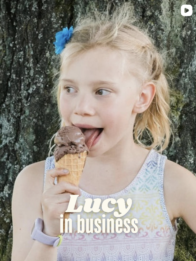A young girl with a blue hair clip licking chocolate ice cream in a cone, standing outdoors in front of a tree. Frozen Custard East Vancouver, BC