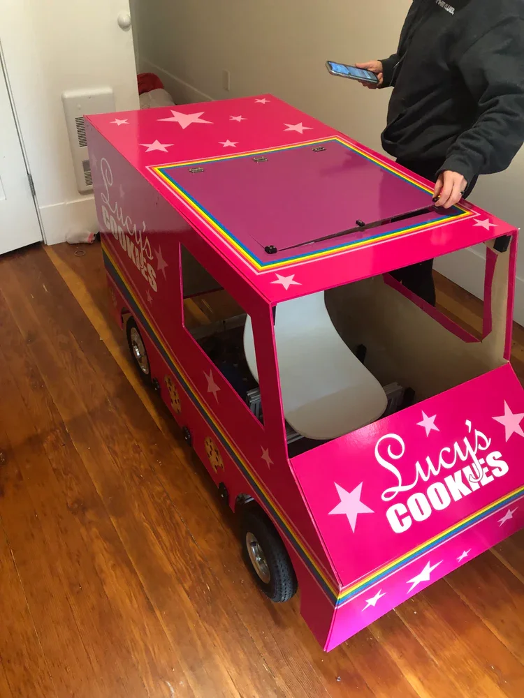 A pink toy car shaped like a delivery vehicle labeled 'Lucy's Cookies,' decorated with stars and rainbow stripes, placed on a hardwood floor with a person standing beside it holding a smartphone. Frozen Custard East Vancouver, BC