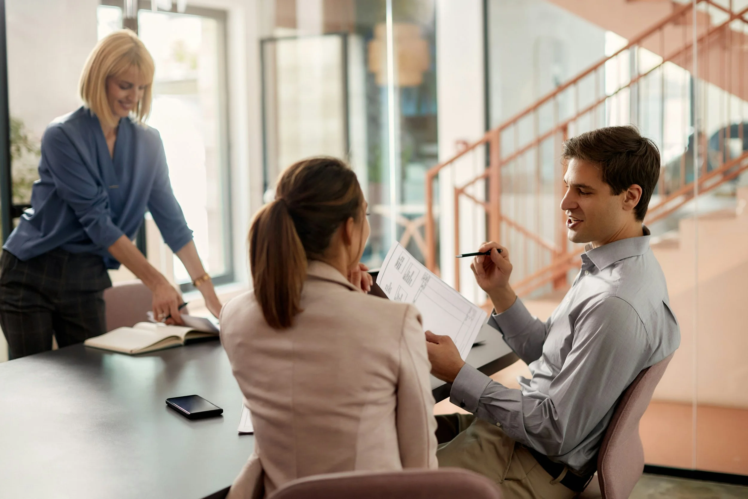 A group of four people in a meeting room, engaging in a discussion. One woman is standing and smiling, while three others sit at the table, one of whom is holding a pen and a document.