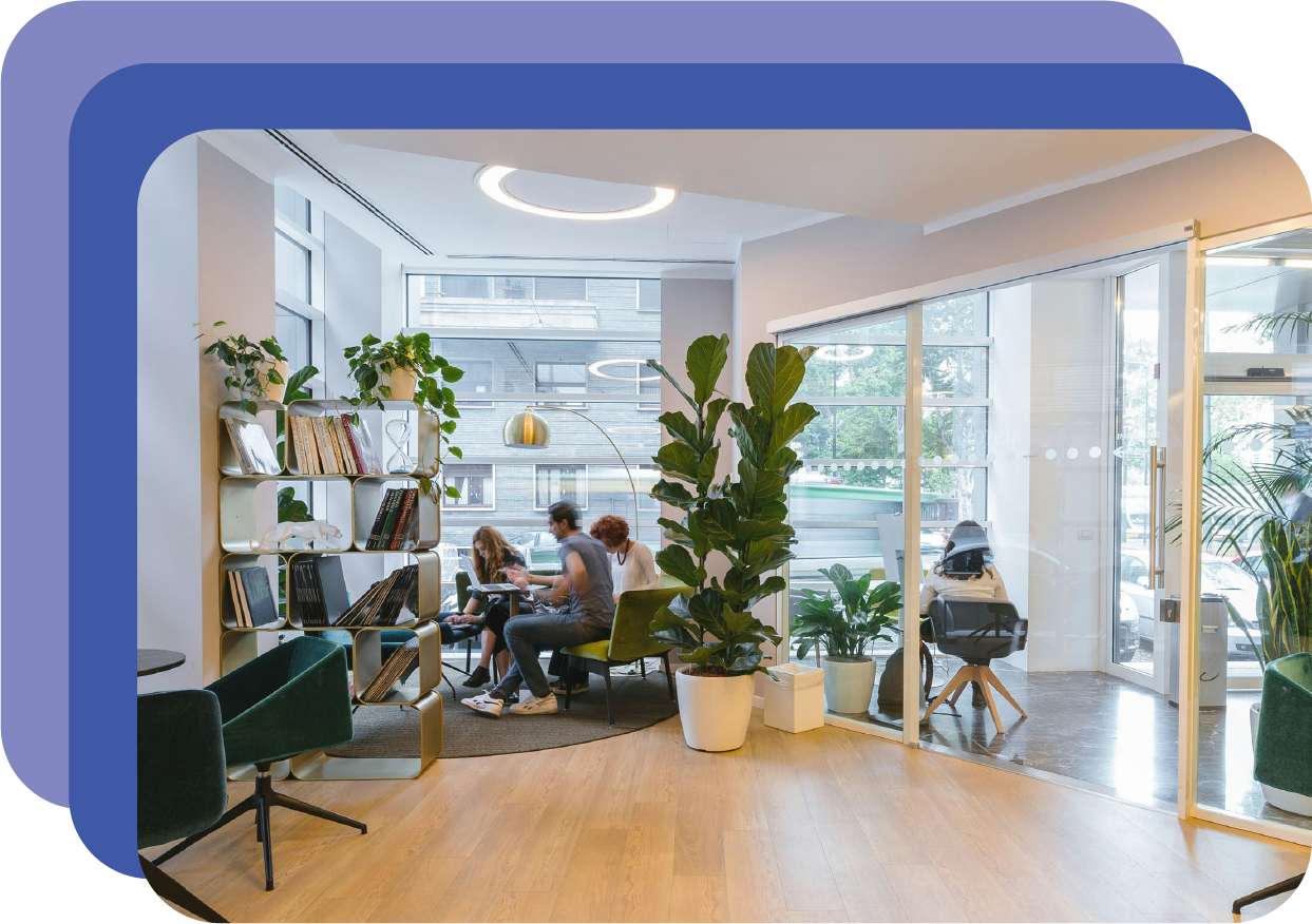 Modern office lounge with large windows, plants, bookshelves, and people working at a table.