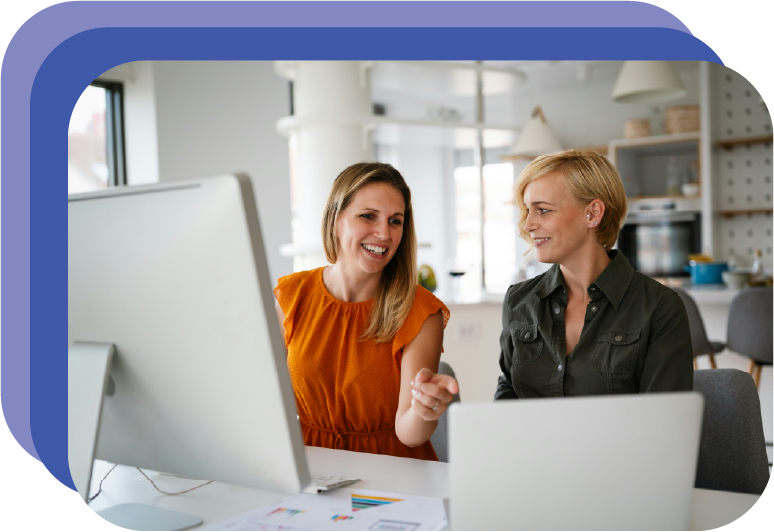 Two women working together at a desk with computer monitors in an office.