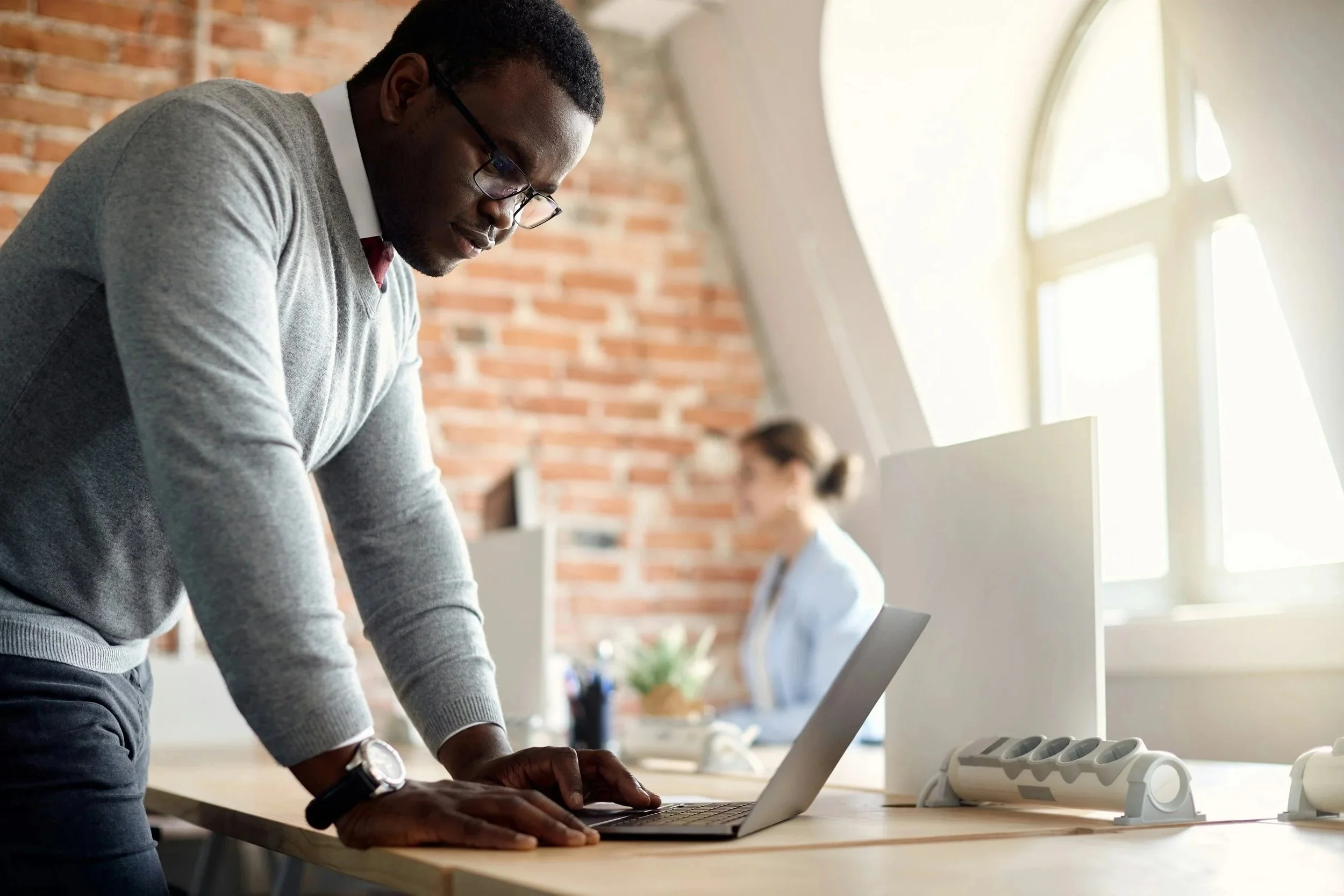 A man wearing glasses, a gray sweater, and a watch, standing at a table, working on a laptop in an office with a brick wall and large windows letting in natural light.