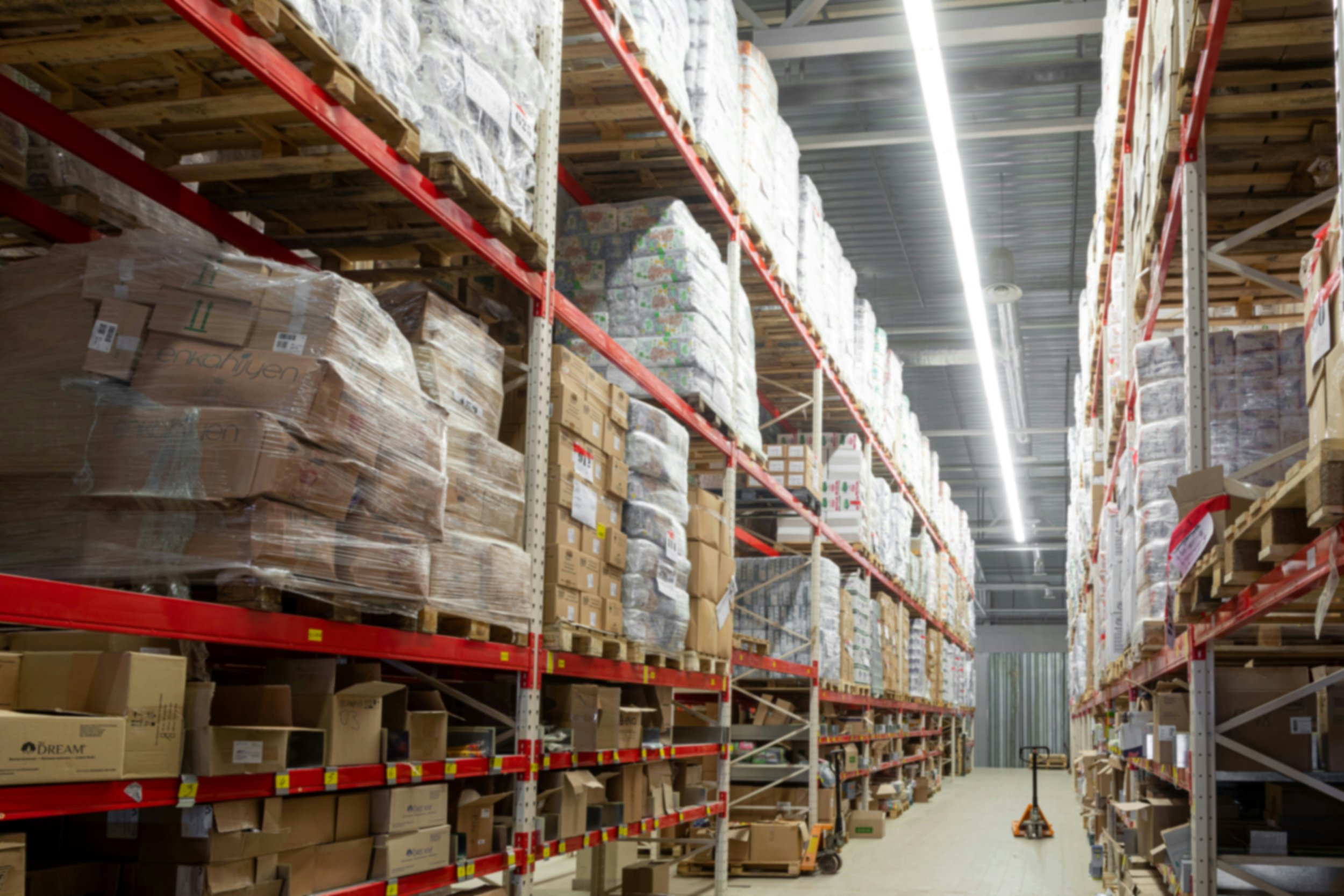 A warehouse aisle with shelves filled with cardboard boxes and wrapped pallets, with a floor buffer machine at the end of the aisle.