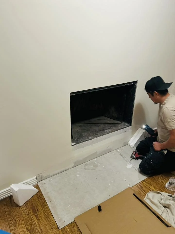 A man kneeling on the floor installing or repairing a fireplace in a room with wooden flooring. The fireplace opening is visible in the wall, and tools and materials are scattered around.