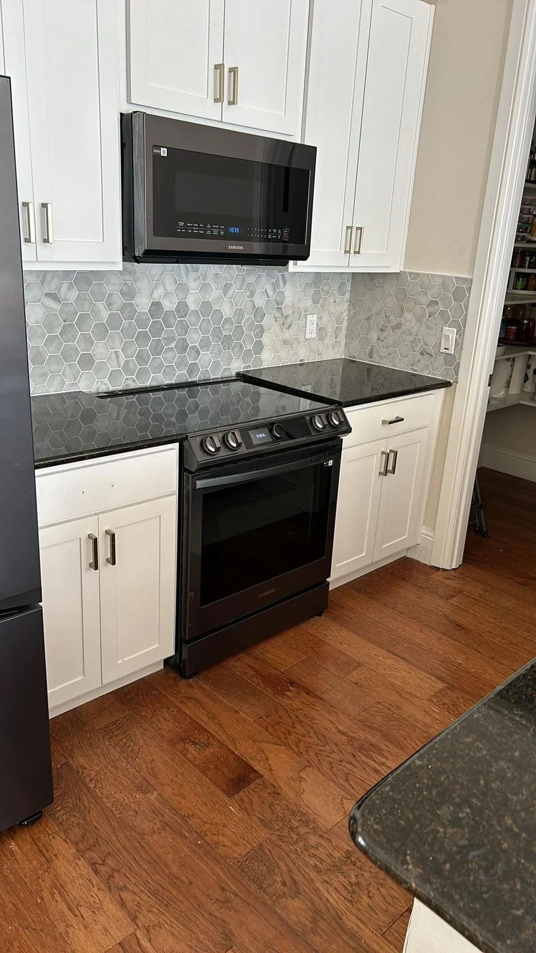 A kitchen with white cabinets, a black stove with oven, and a black microwave mounted above the stove. The backsplash is made of gray hexagonal tiles, and the floor is wood. A corner of a black granite countertop is visible in the foreground.