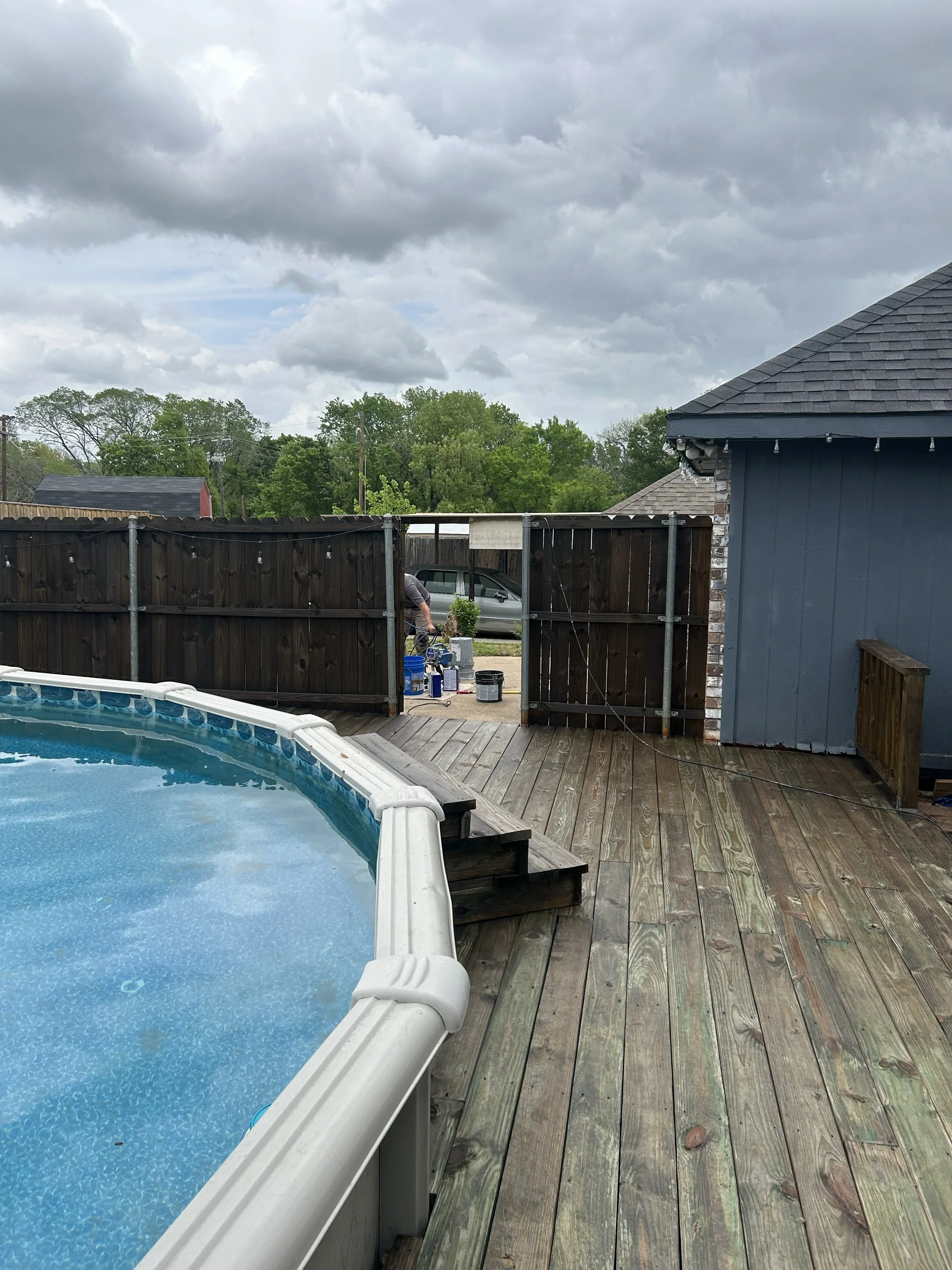 Backyard with a swimming pool, wooden deck, gray fence, and cloudy sky overhead. A person is working near the fence, with tools and paint buckets visible.