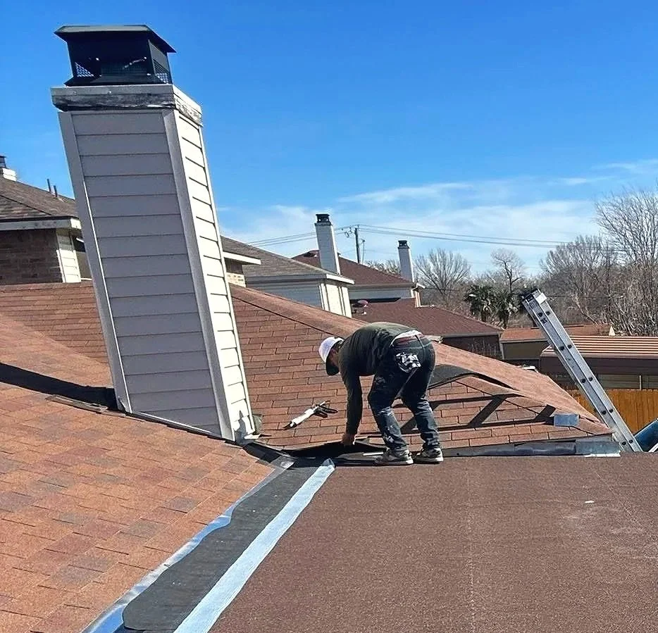 A person working on a roof with shingles, a chimney, and a ladder, under a clear blue sky.