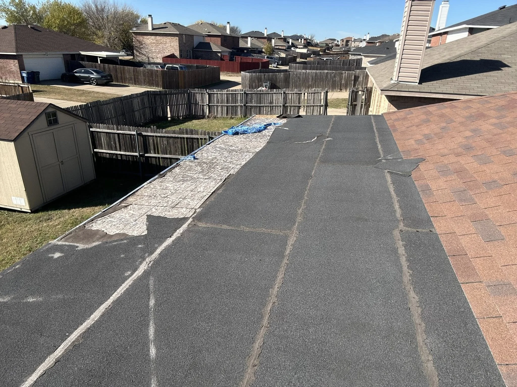 View from a rooftop showing neighboring houses, fences, a small yard, and parked cars on a street in a suburban neighborhood on a sunny day.