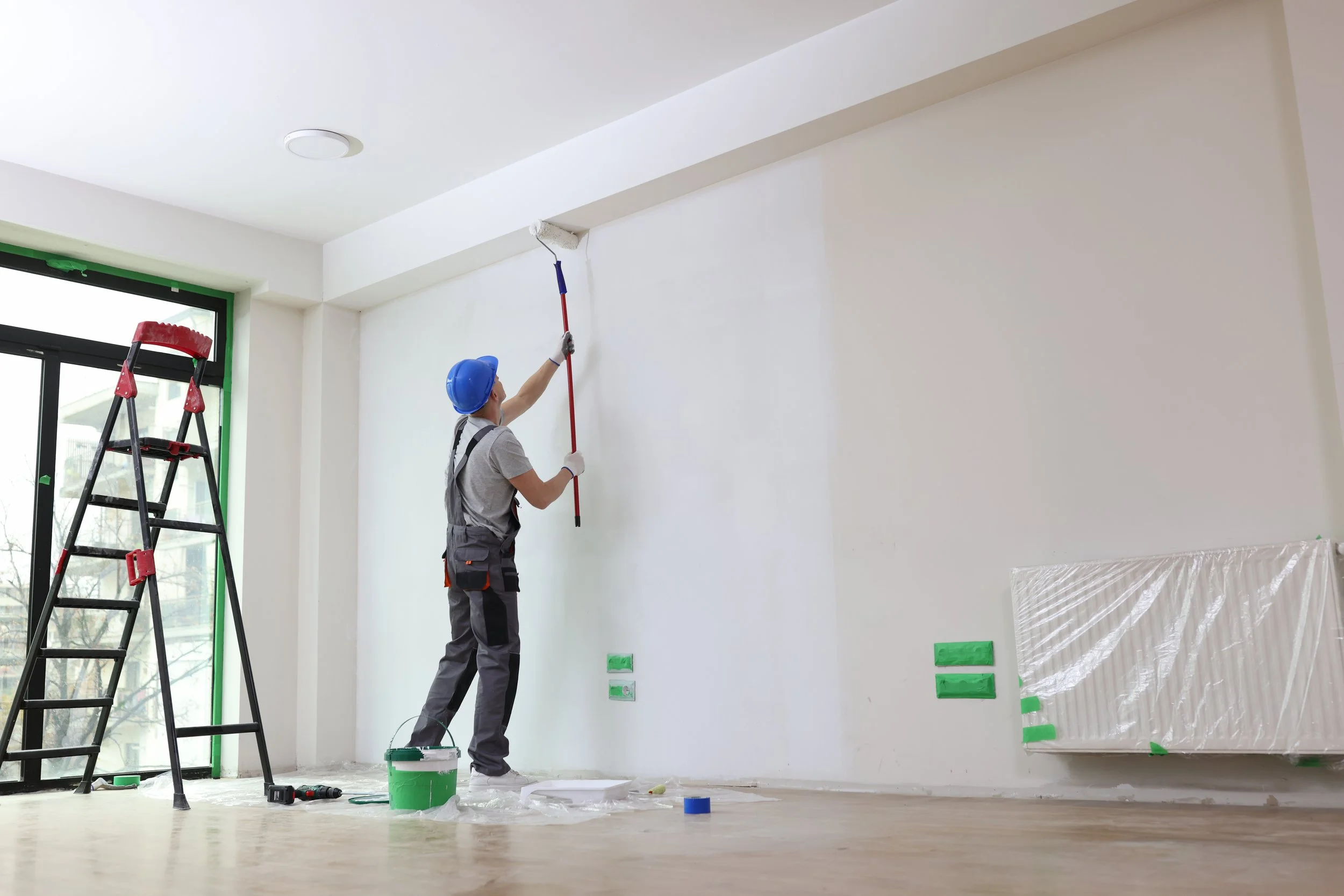 A person wearing a blue safety helmet and grey work clothes is painting a white wall with a roller brush. There is a black ladder with red accents near a large window, and a bucket of white paint on the floor. The room appears to be under renovation, with plastic coverings taped to the wall and floor.