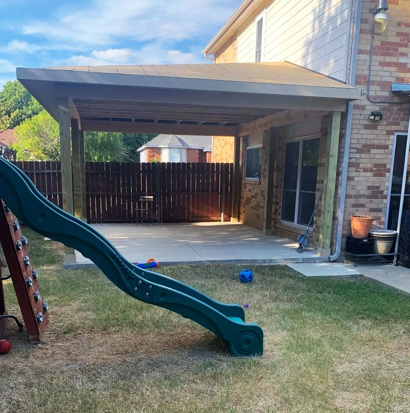 Backyard patio with a sliding glass door, a wooden overhang, a children's slide, toys on grass, and a fenced yard with trees