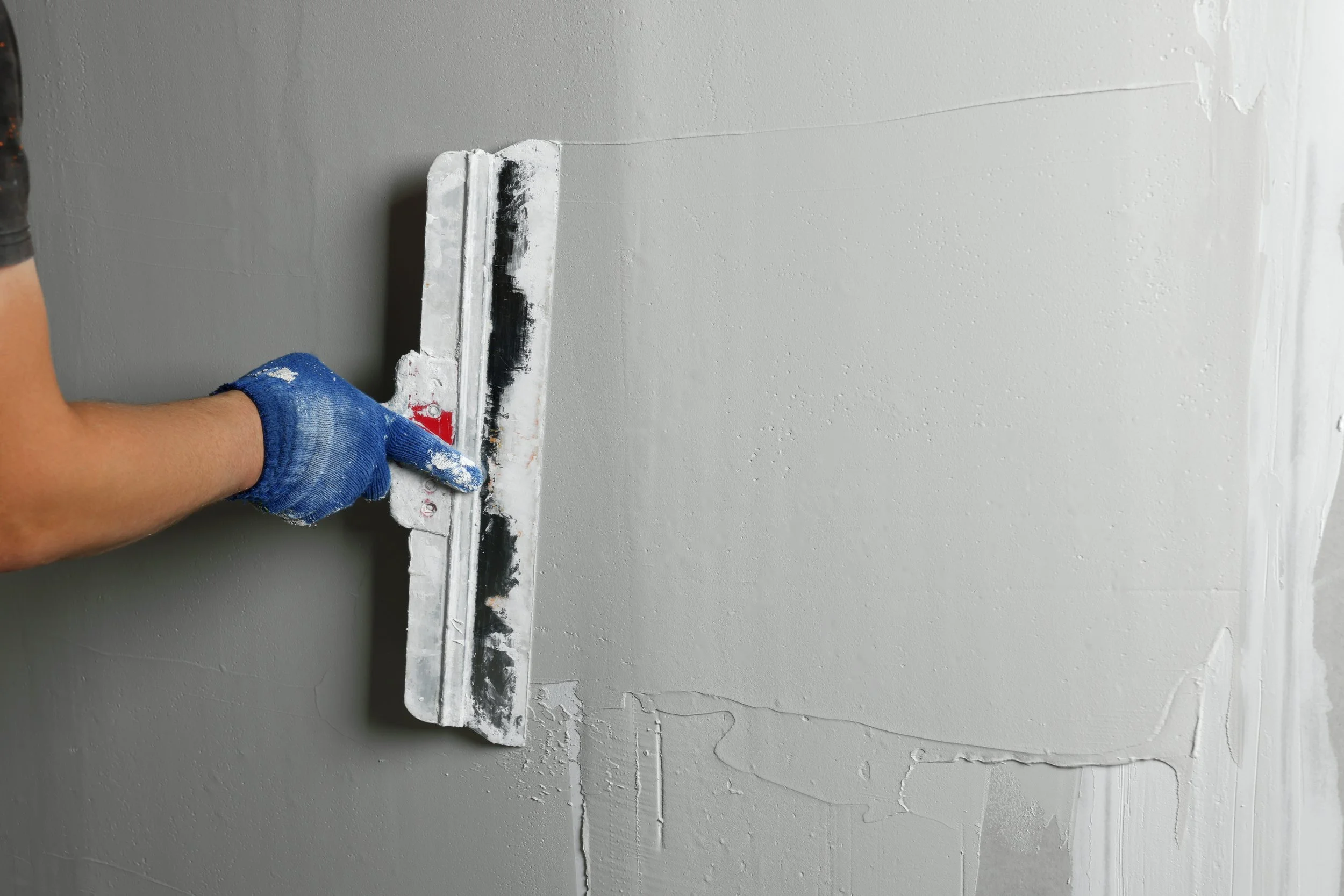 Person wearing a blue glove smoothing fresh white plaster on a wall with a putty knife.