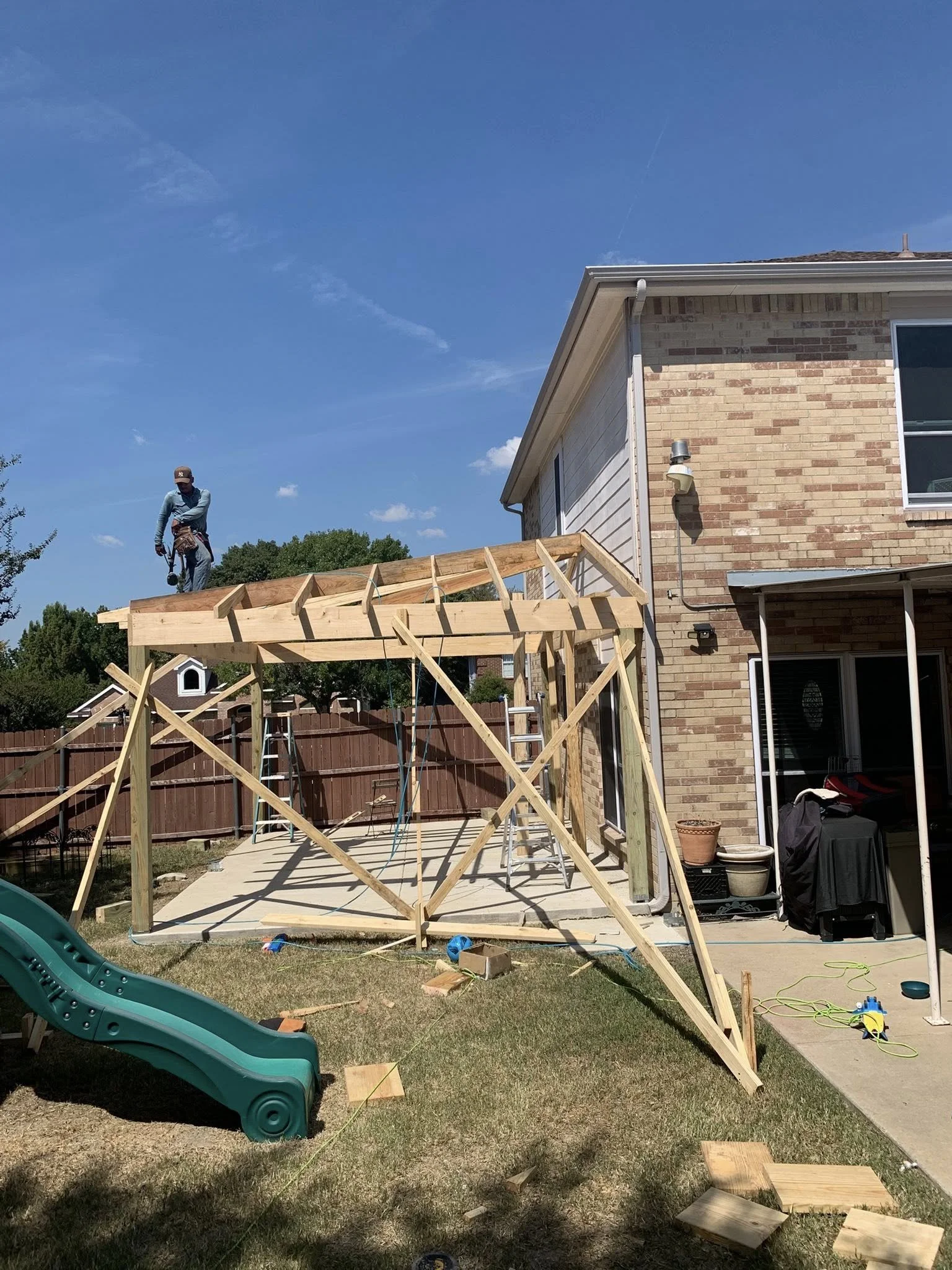 A person standing on a wooden porch frame under construction next to a brick house, with construction tools and materials on the ground and a green slide in the yard.