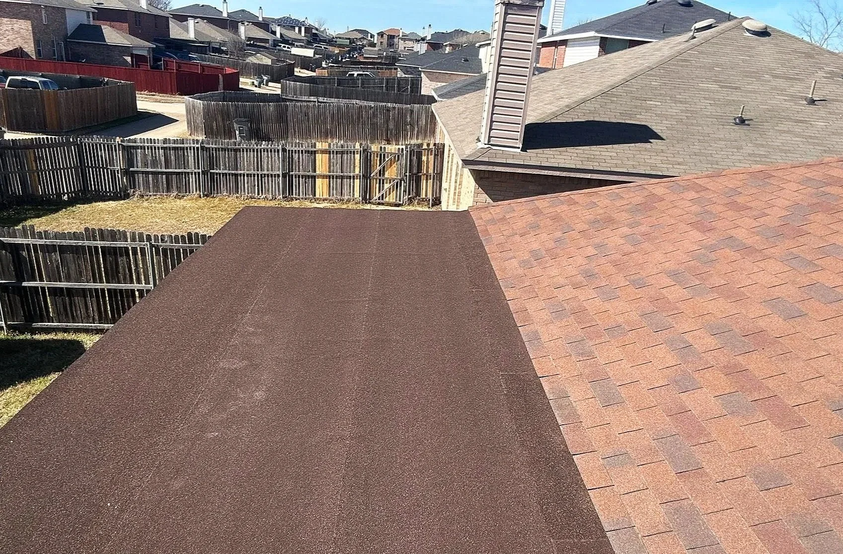 View from a rooftop showing the neighboring rooftops, fences, and yards in a residential area with clear skies.