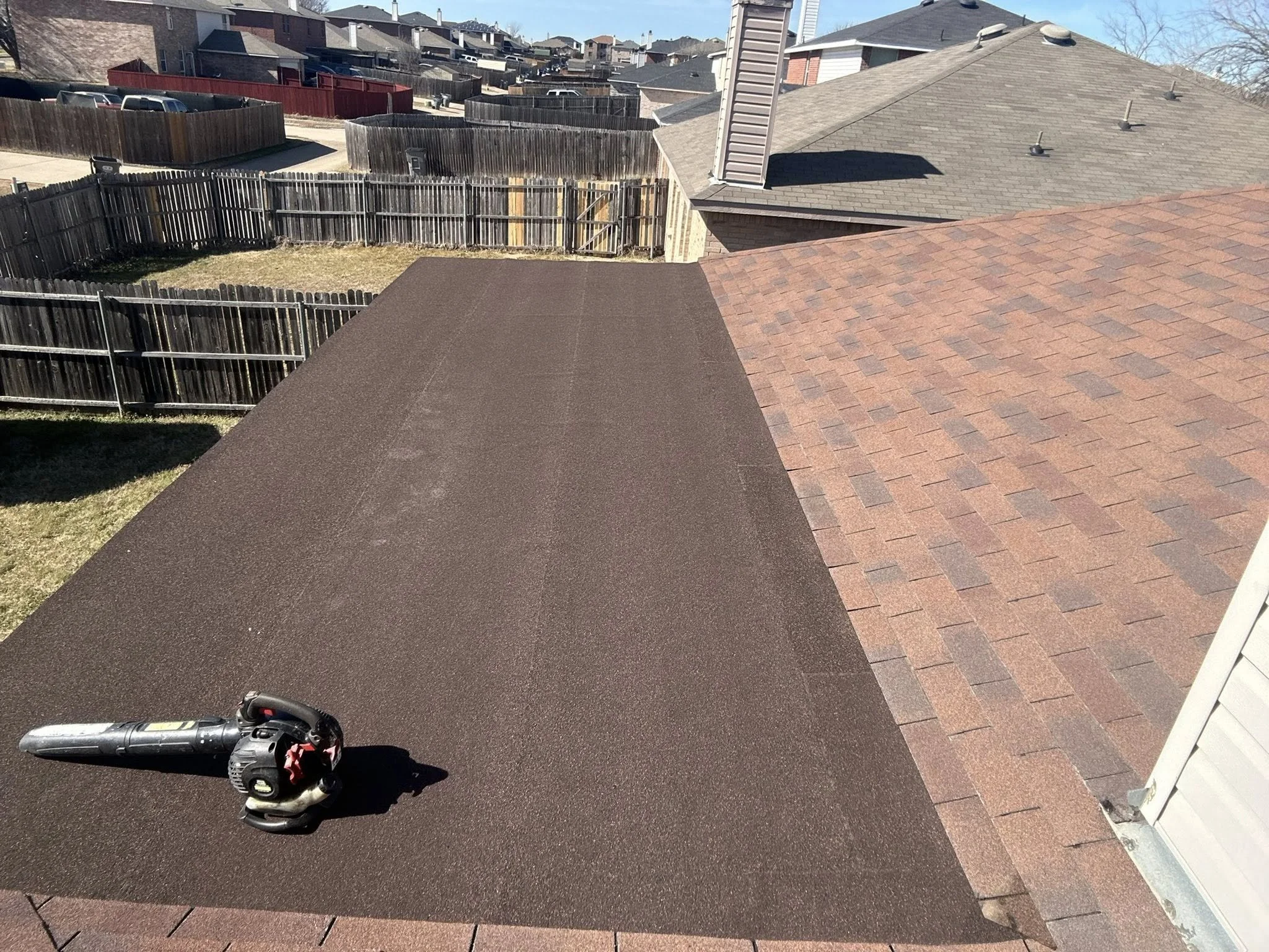 A backyard with a brown textured driveway, a fence, a brick house roof with a chimney, and a black leaf blower on the driveway.