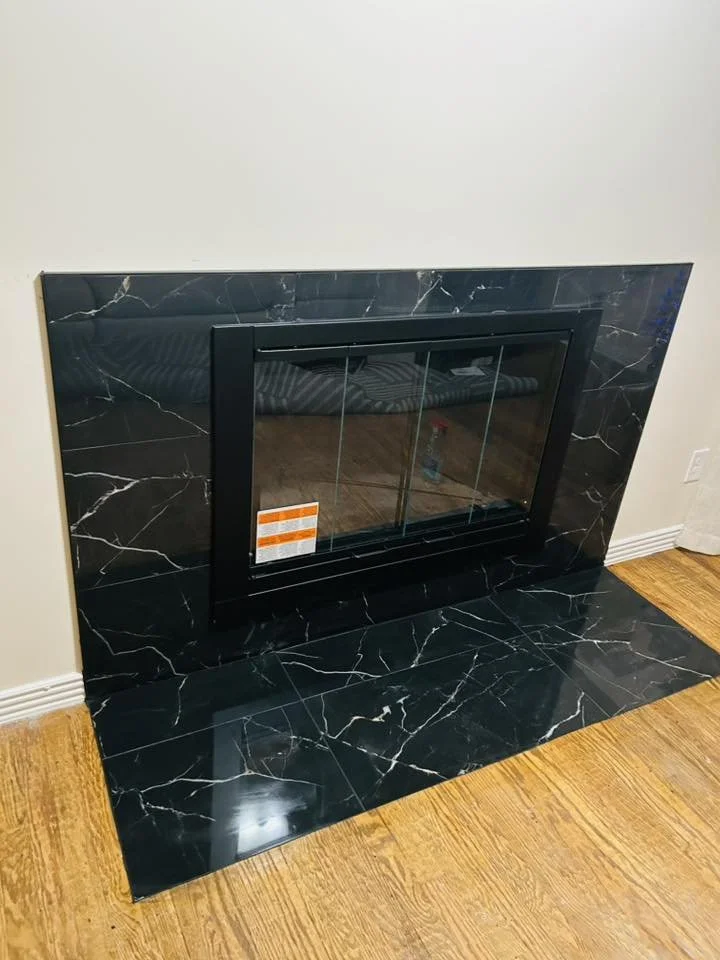 Black marble fireplace with glass doors and a black frame, installed on a wall with a wood floor nearby.