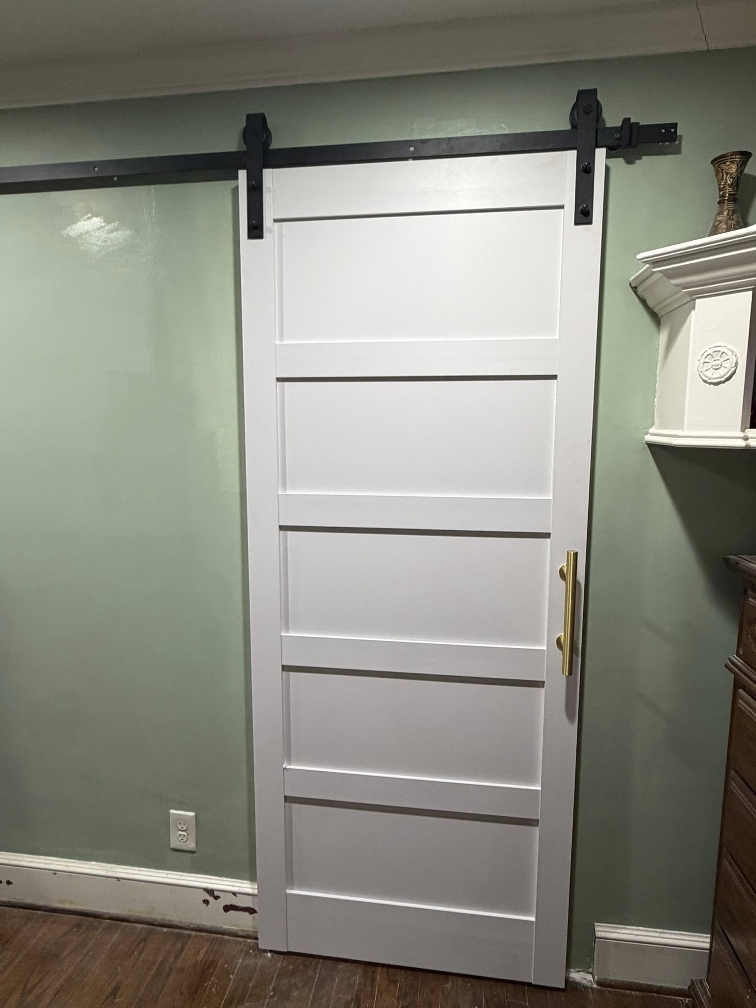 A white sliding barn door with horizontal panels and a gold handle, mounted on a black metal track, installed on a green wall.