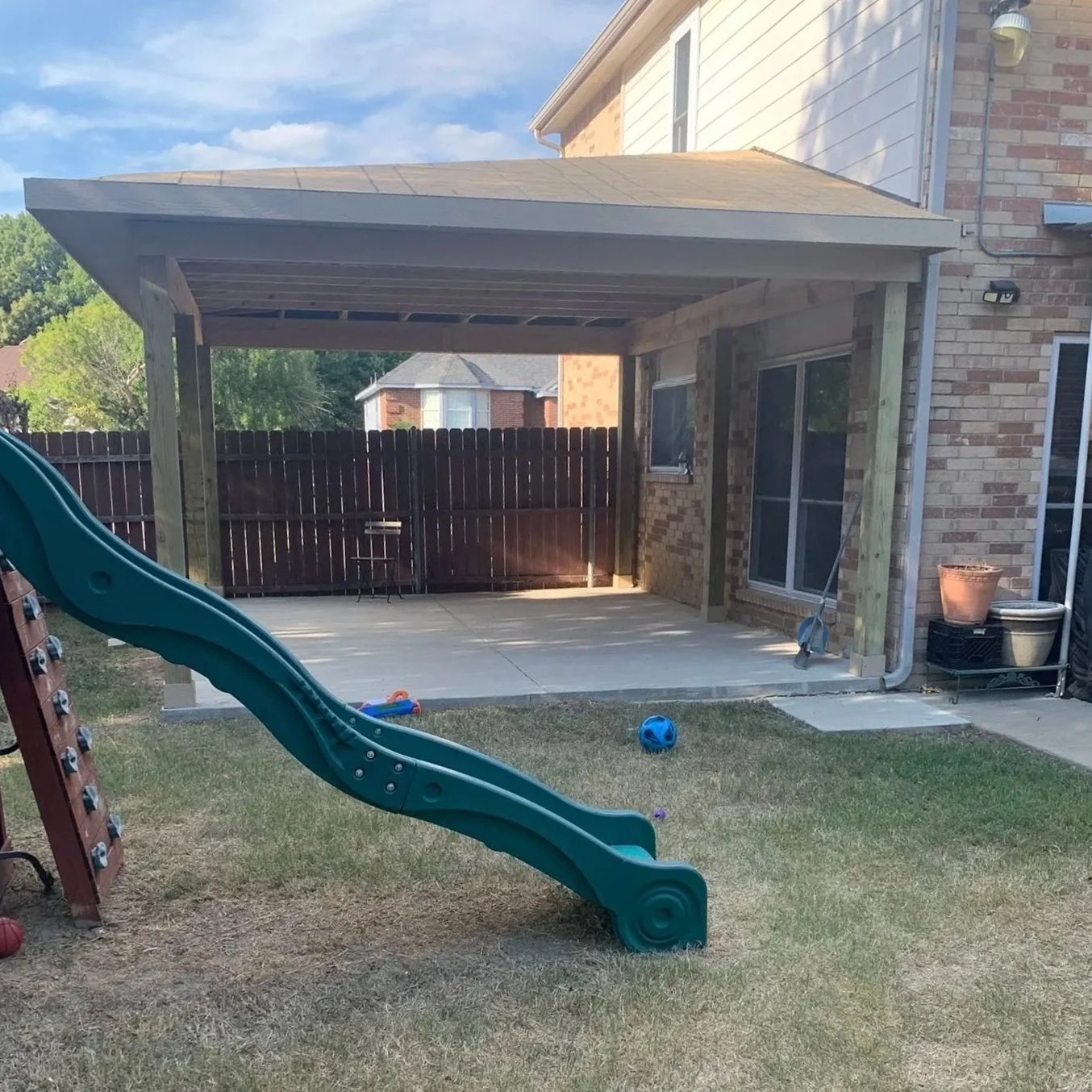 Backyard with a covered patio area, children's slide, and toys on the grass. Fence and neighboring houses in the background.