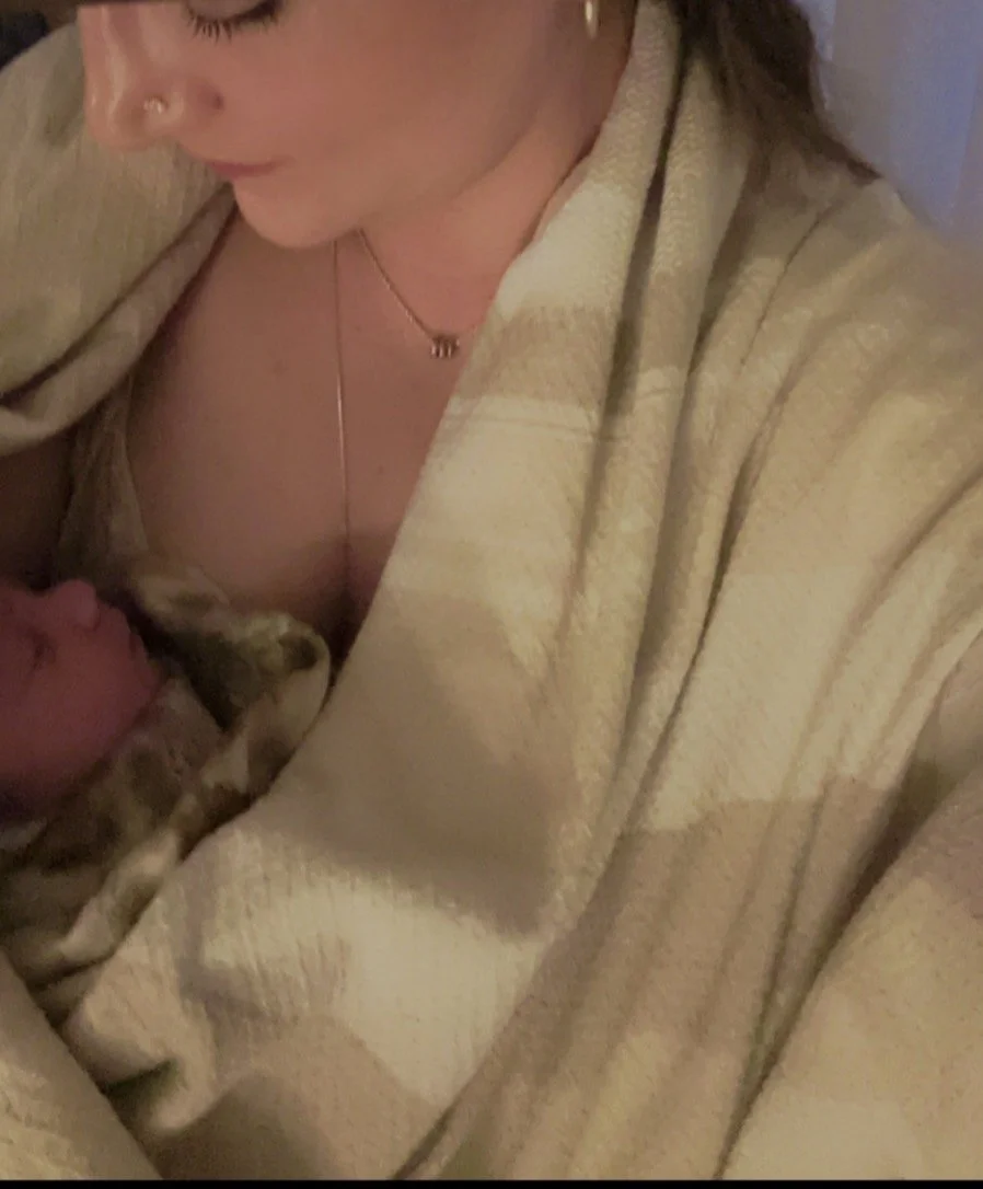 A woman holding a sleeping kitten wrapped in a blanket, near her chest, with a neutral-colored blanket draped over her shoulders.
