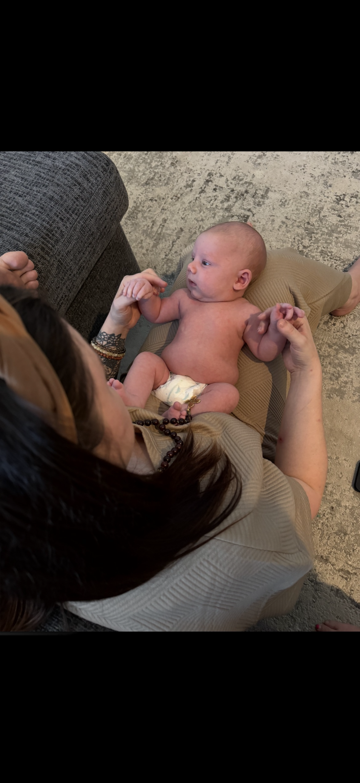 A woman lying on her back holding a newborn baby on her lap, with the baby's arms and legs raised, in a cozy indoor setting.